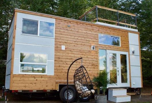 casa sobre rodas de madeira com detalhes brancos na porta e janelas, além de um espaço em cima da casa (como uma varanda(