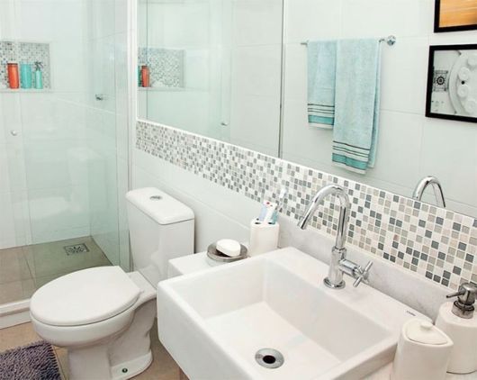 Banheiro com louças brancas, faixa de pastilhas em vários tons de cinza e grande espelho horizontal sobre a pia