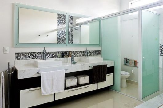 banheiro com box de vidro, faixa de pastilhas em cores preta, cinza e branca sobre a pia de louça branca
