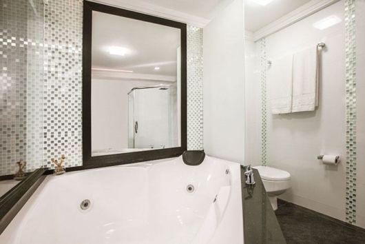 banheiro com louças brancas, grande espelho com molde preto e faixa de pastilhas na vertical em tons de cinza com branco