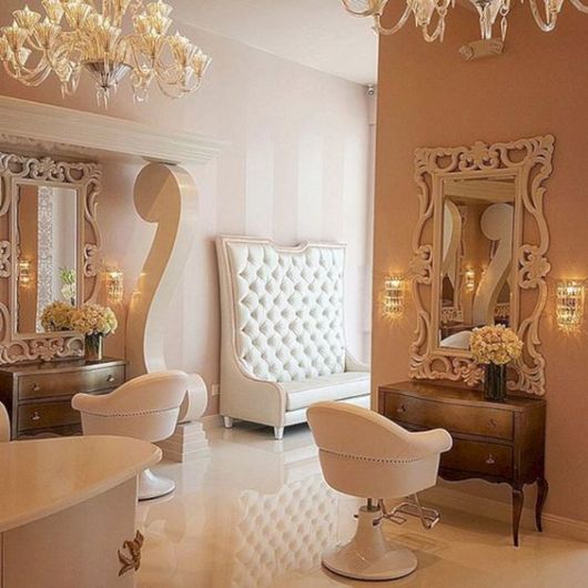 salão de beleza com parede em tons de rosa claro, móveis brancos e grandes lustres de cristal pendentes. Sofá com espalda alta ao fundo