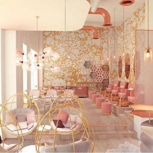 salão de beleza com papel de parede branco e dourado, cadeiras com espaldas douradas e ao fundo cadeiras em tom de rosa claro