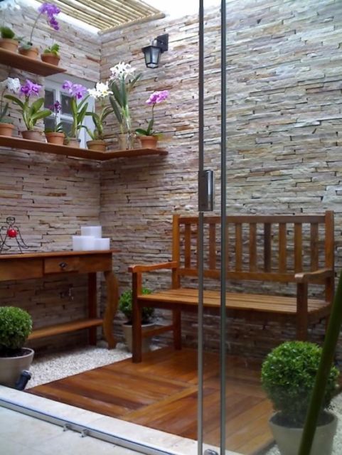 jardim interno com parede recoberta de pedra, banco de madeira e vasos de flores e plantas