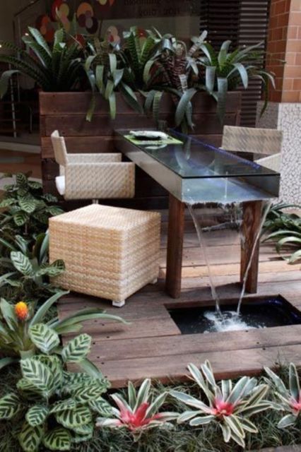 mesa de vidro com cascata de água perpassando seu interior e vasos de plantas ao fundo