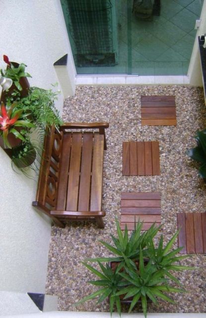 Jardim interno com chão recoberto de pedras marrons, banco de madeira e porta de vidro, além de vasos de plantas