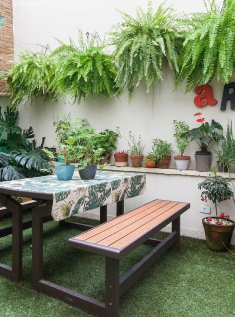 Esepaço com parede branca, samambaias penduradas na parede, mesa retangular com toalha de folhagens e muitos vasos de plantas