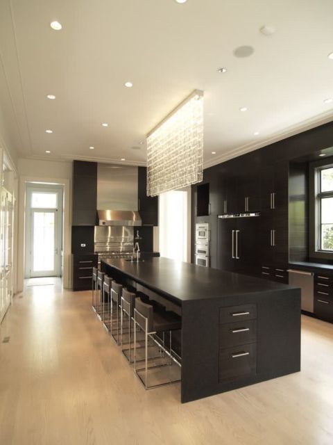 cozinha com ilha e armários pretos, piso de madeira, teto branco com grande lustre de cristal disposto na horizontal pendente do teto