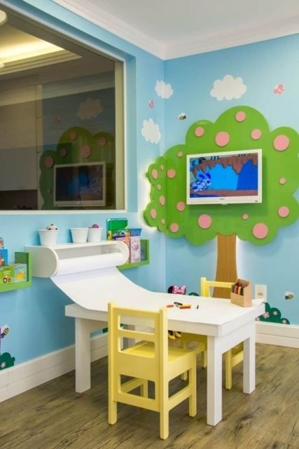 sala para crianças com paredes azuis, árvore verde pintada na parede, pequena mesa branca com cadeiras amarelas e piso de madeira