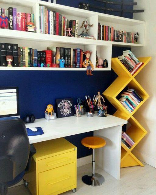 área de escritório com parede azul escuro, móveis amarelo vivo, estante suspensa com nichos branca e muitos livros e alguns bonecos personagens de filmes 