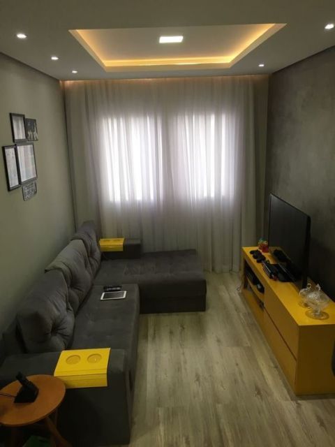 equena sala com piso de madeira, sofá cinza escuro com chaise, janela ao fundo e televisão sobre móvel de madeira